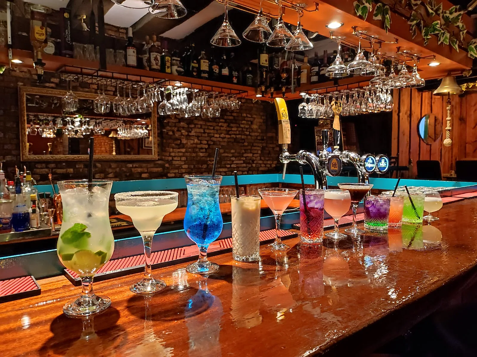 Een rij cocktails op de mahoniehouten bar, met hangende glazen en de taps van Leffe en Hertog Jan