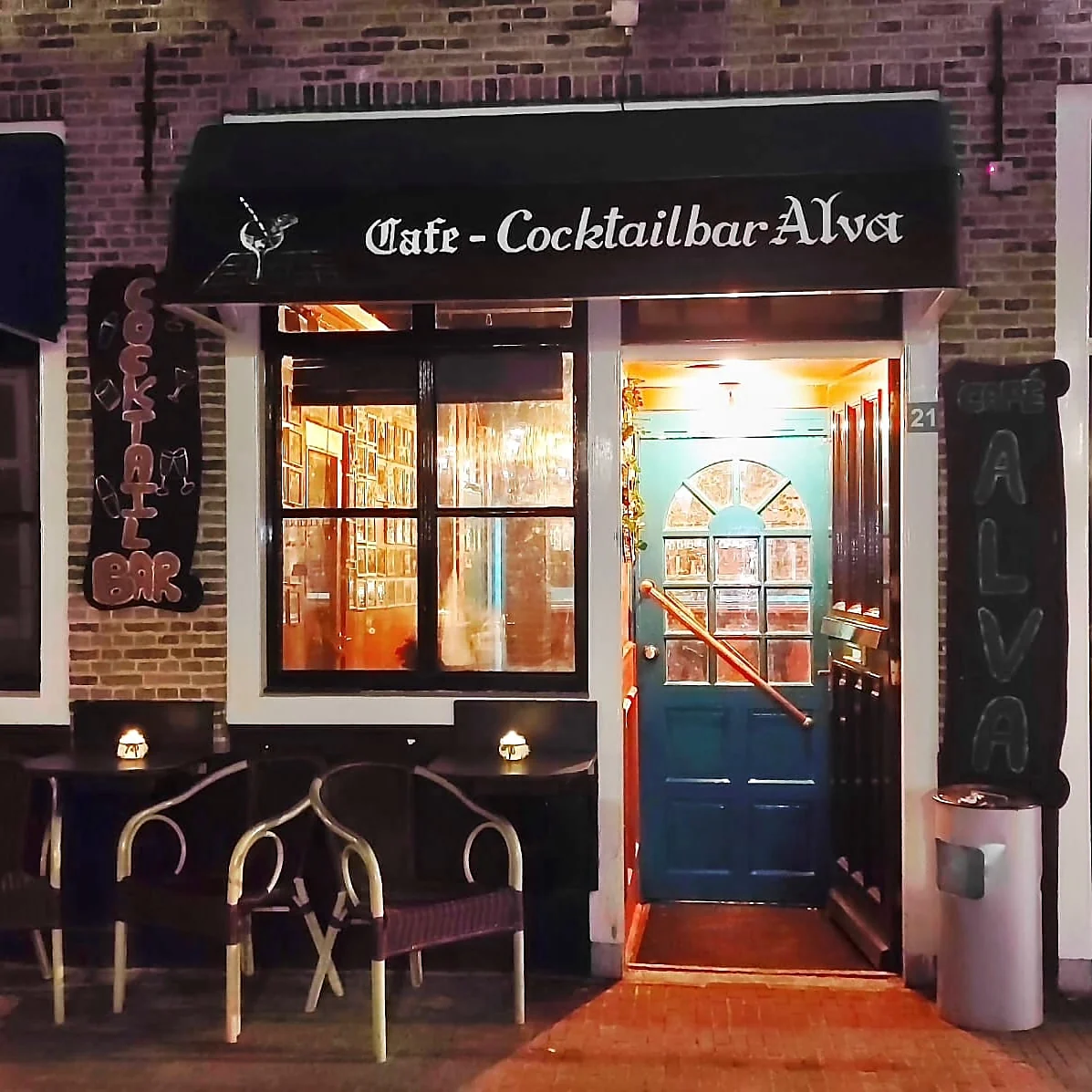 De voorgevel van Café-Cocktailbar Alva 's avonds: zwarte luifel, verlichte ramen en de groenblauwe voordeur